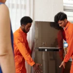 Two men in matching orange jumpsuits carefully lift and move a dark refrigerator inside a bright room, with the blurry shoulder of another person in a blue top in the foreground.