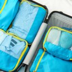 A flat lay, overhead view of an open black suitcase packed with blue and gray clothing, a blue cycling helmet, black and neon green athletic shoes, espadrilles, a camera, and a wallet. A person's hands are placing a light blue collared shirt into the right side.