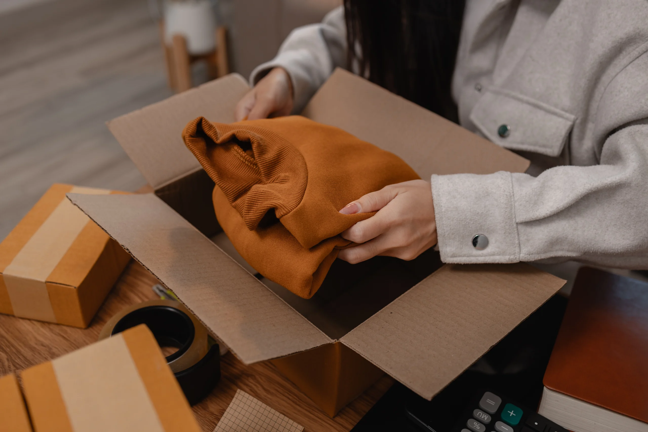 A person's hands wearing a light gray jacket placing a folded, orange-brown sweatshirt or garment into an open cardboard box on a wooden desk with other boxes and a calculator visible.